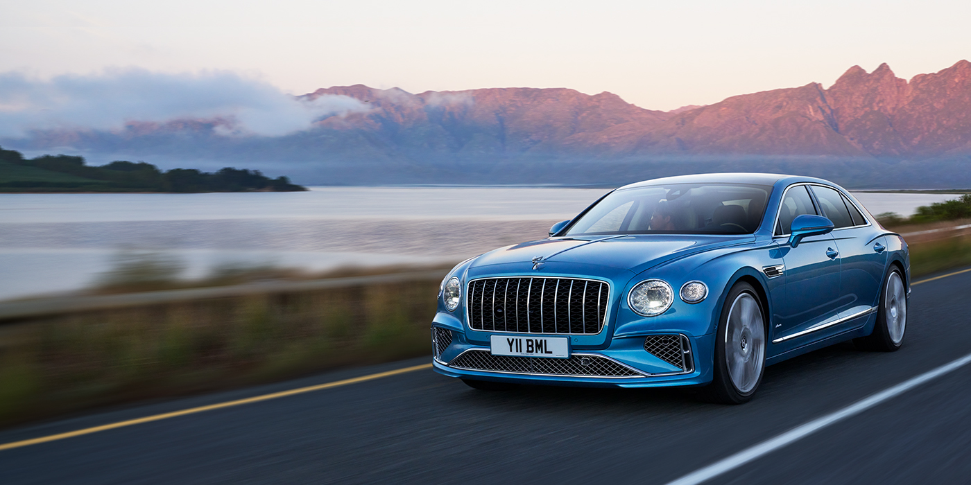 Exclusive Cars Vertriebs GmbH New Bentley Flying Spur Azure sedan in Light windsor blue paint by Mulliner, with 22 inch Azure wheels and chrome straight vaned grille driving on a mountain road