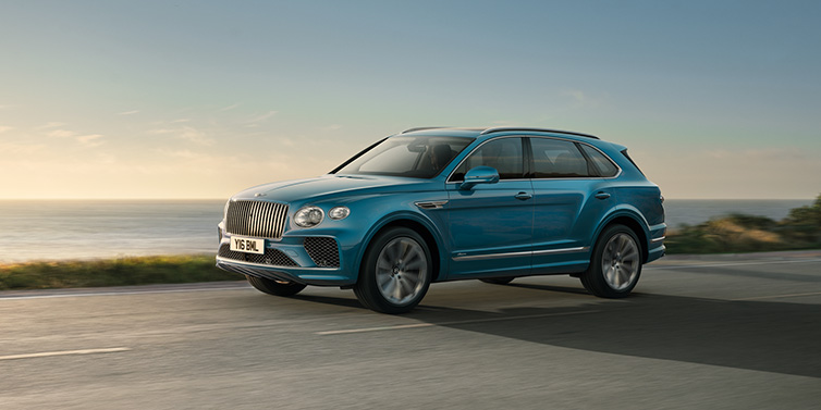 Exclusive Cars Vertriebs GmbH Bentley Bentayga Azure SUV in Topaz blue paint driving dynamically by the ocean with 22 inch 10 spoke directional wheels