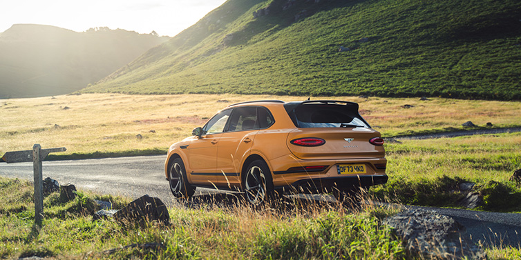 Exclusive Cars Vertriebs GmbH Bentley Bentayga S SUV rear three quarter in Sunburst Gold paint on a road surrounded by green hills